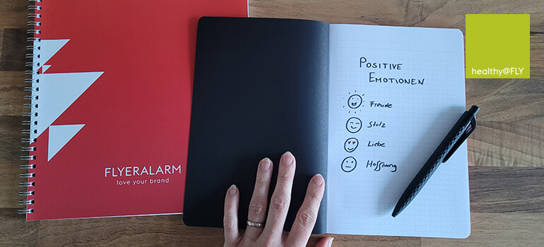 Auf einem Tisch liegen zwei Notizbücher, eines offen mit einer Liste positiver Emotionen wie Freude und Mut.