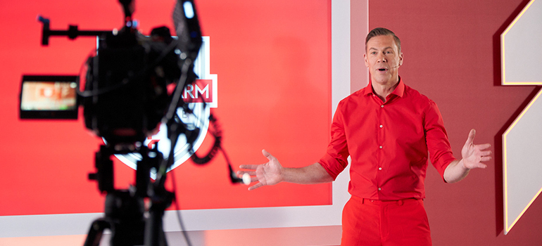 Mann in rotem Outfit spricht vor Kamera in TV-Studio mit FLYERALARM-Logo.